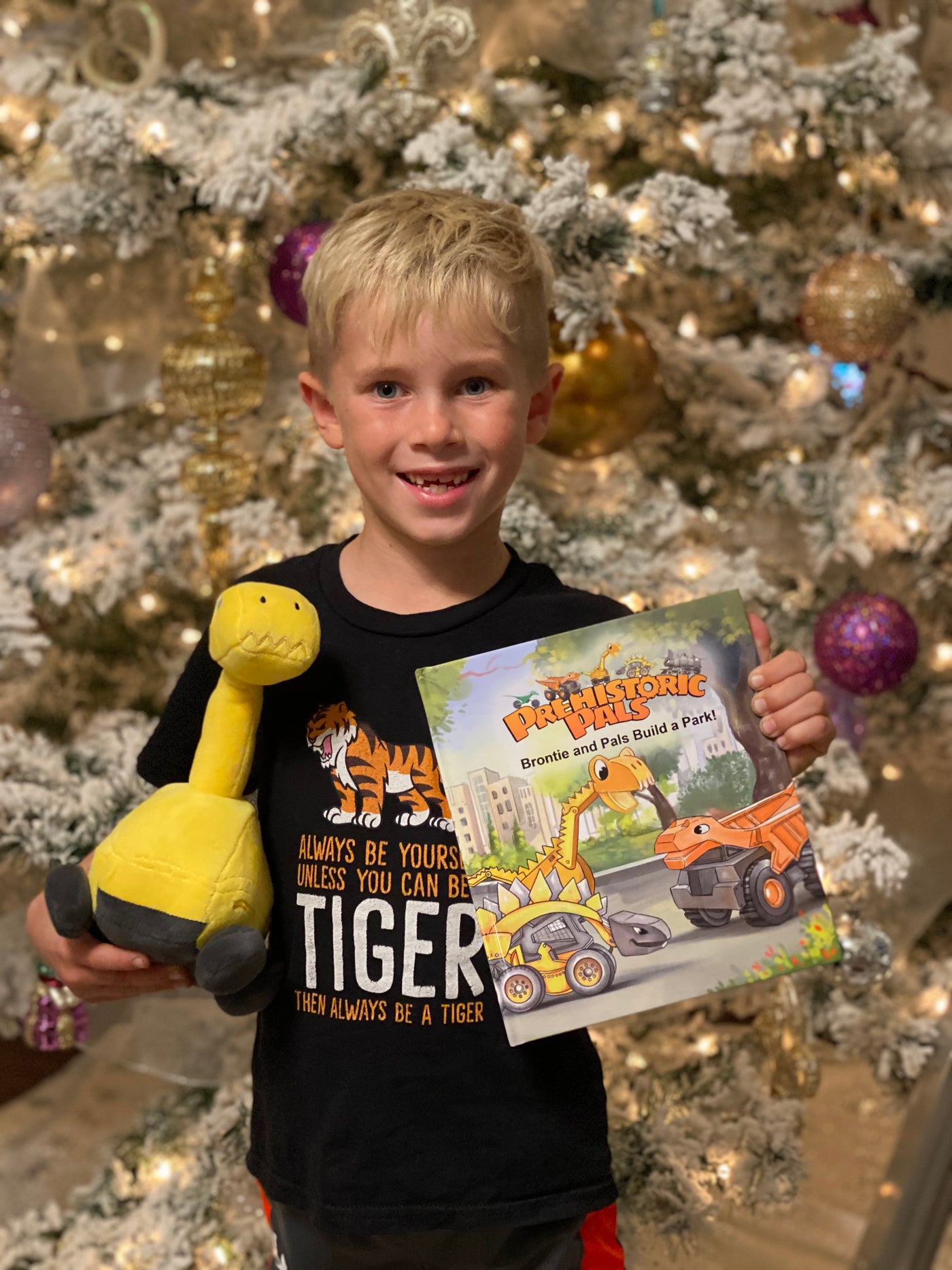 A 6 year old boy is holding a plush toy Brontie. He also has the prehistoric pals toys book, Brontie and Pals Build a Park! book in the other hand. The little boy is standing in front of a decorated Christmas tree.